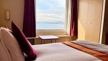 A hotel room with a bed, a small table, a view of a city skyline through a window, and a door.