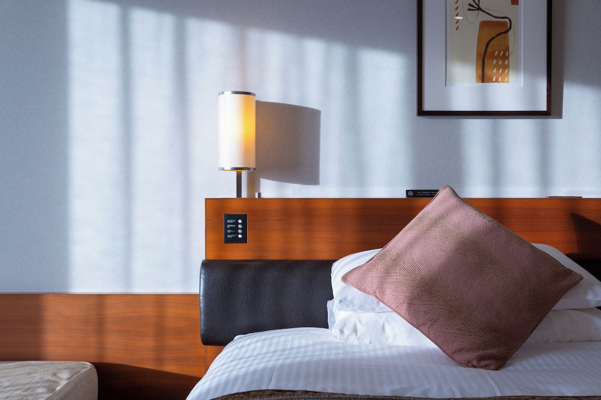 A hotel room with a bed, a bedside lamp, a control panel, and a framed artwork on the wall.