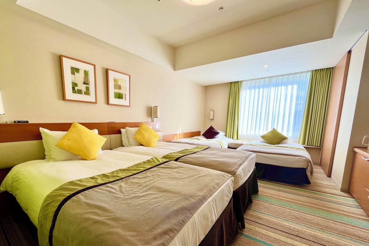 A hotel room with three beds, each with a different colored bedspread. There are two framed pictures on the wall, a window with curtains, and a small desk with a lamp.