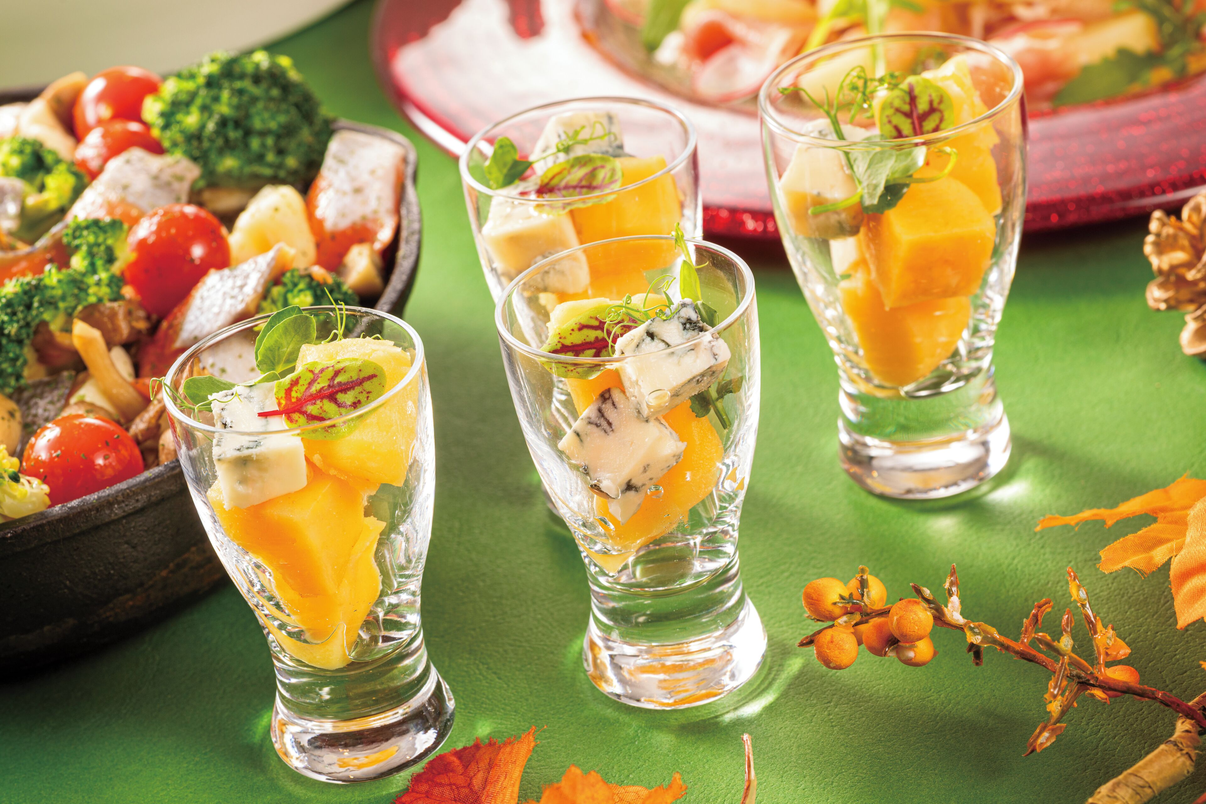 Three glasses of a fruit-based drink with garnishes, a bowl of mixed vegetables, and some decorative elements on a green surface.
