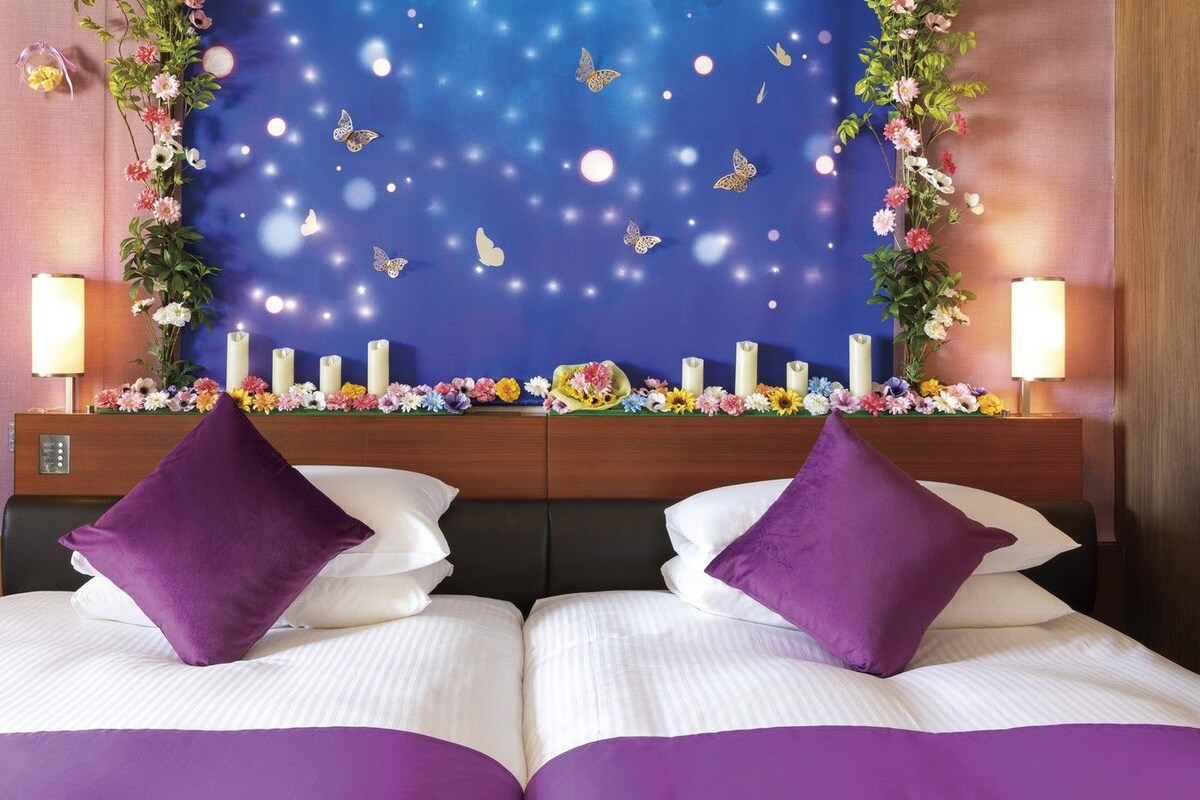 A hotel room with a bed, two purple pillows, a nightstand with a lamp, a wall with a decorative tapestry, and a floral arrangement.