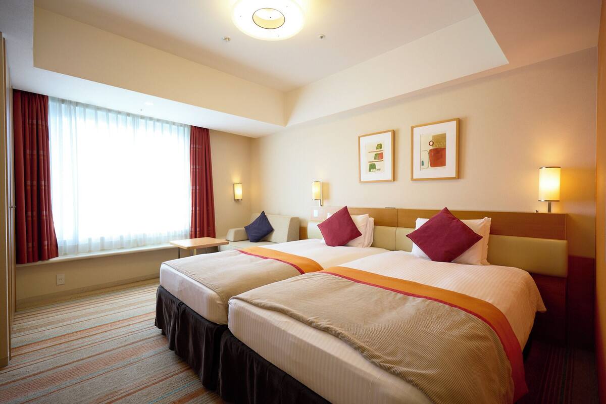 A hotel room with two beds, a window with curtains, wall-mounted lamps, and framed artwork.
