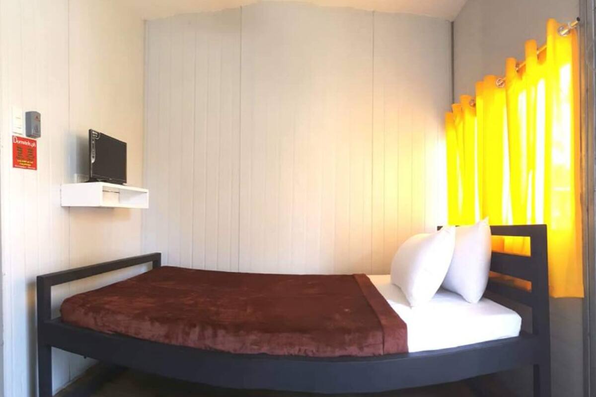 A single bed with a headboard, a small shelf with a television, and a window with yellow curtains.