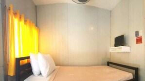 Ein Einzelbett mit weißer Matratze und Kissen, ein kleiner Nachttisch, ein Fernseher auf einem Regal und ein Fenster mit gelben Vorhängen.