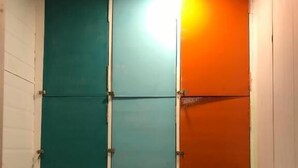 Ein schmaler Korridor mit türkisfarbenen und orangefarbenen Wänden, einer weißen Tür links und einer Leuchte an der Decke.