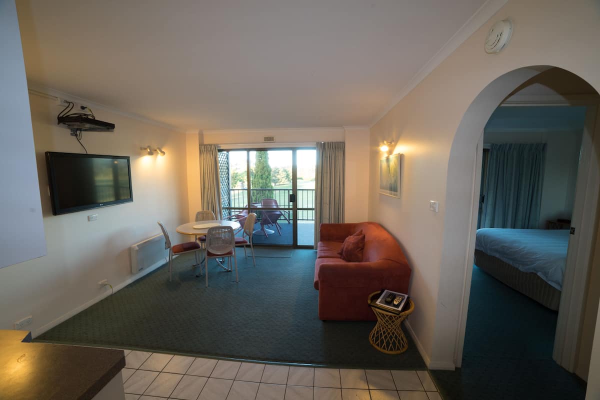 A hotel room with a red sofa, a TV mounted on the wall, a dining area with a table and chairs, and a balcony with a view of trees.