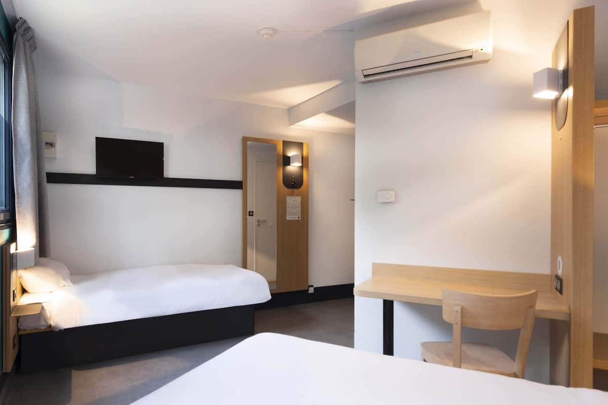 A hotel room with two beds, a desk, a chair, a TV, and an air conditioning unit.