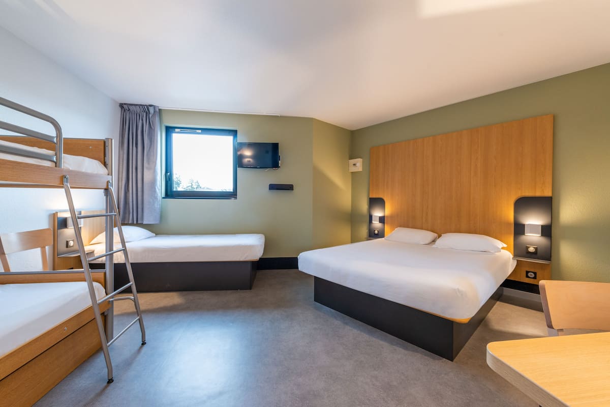 A hotel room with a bunk bed, a single bed, a desk, a chair, a TV, and a window.
