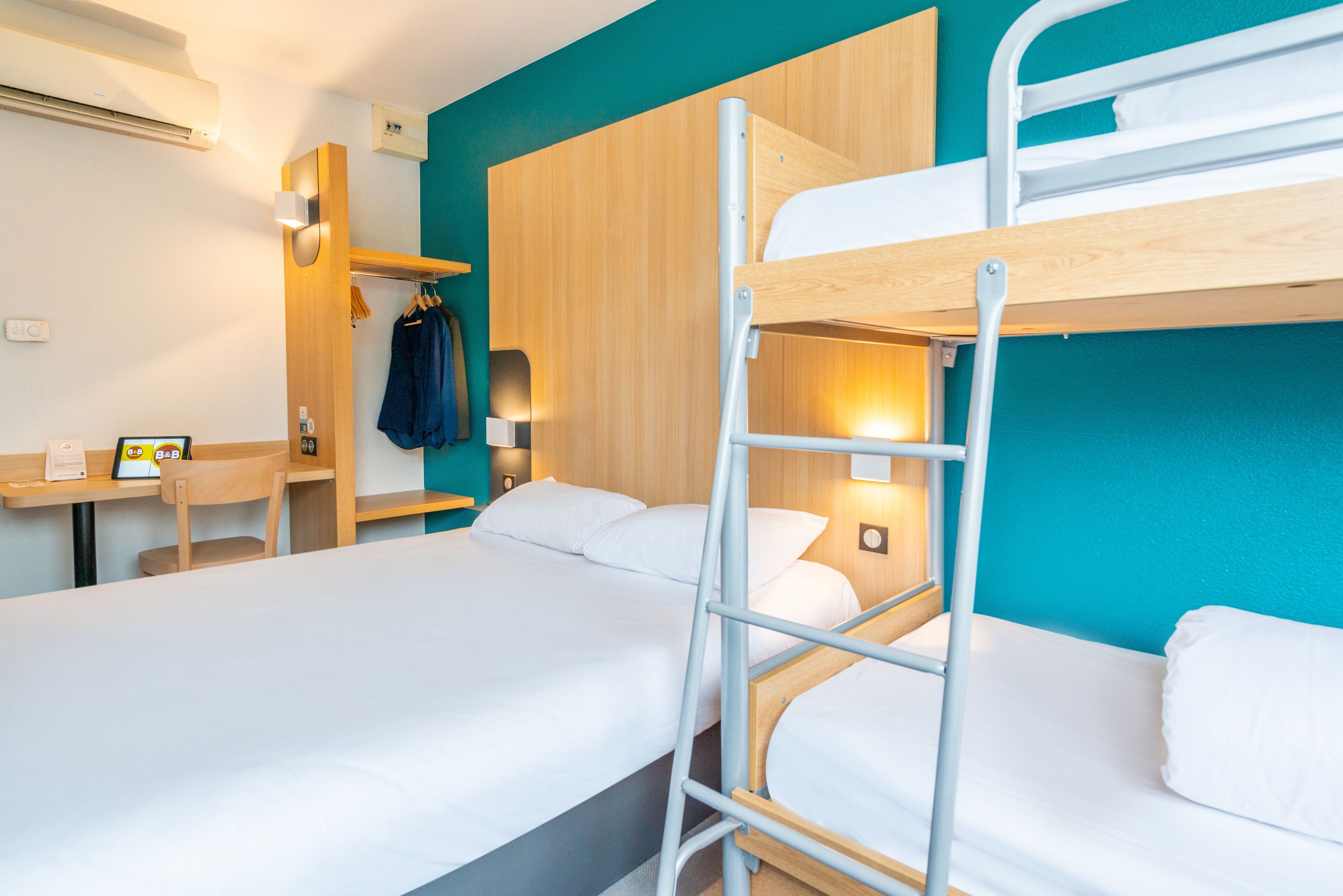 A hotel room with bunk beds, a desk with a computer, and a wall-mounted shelf with clothes.