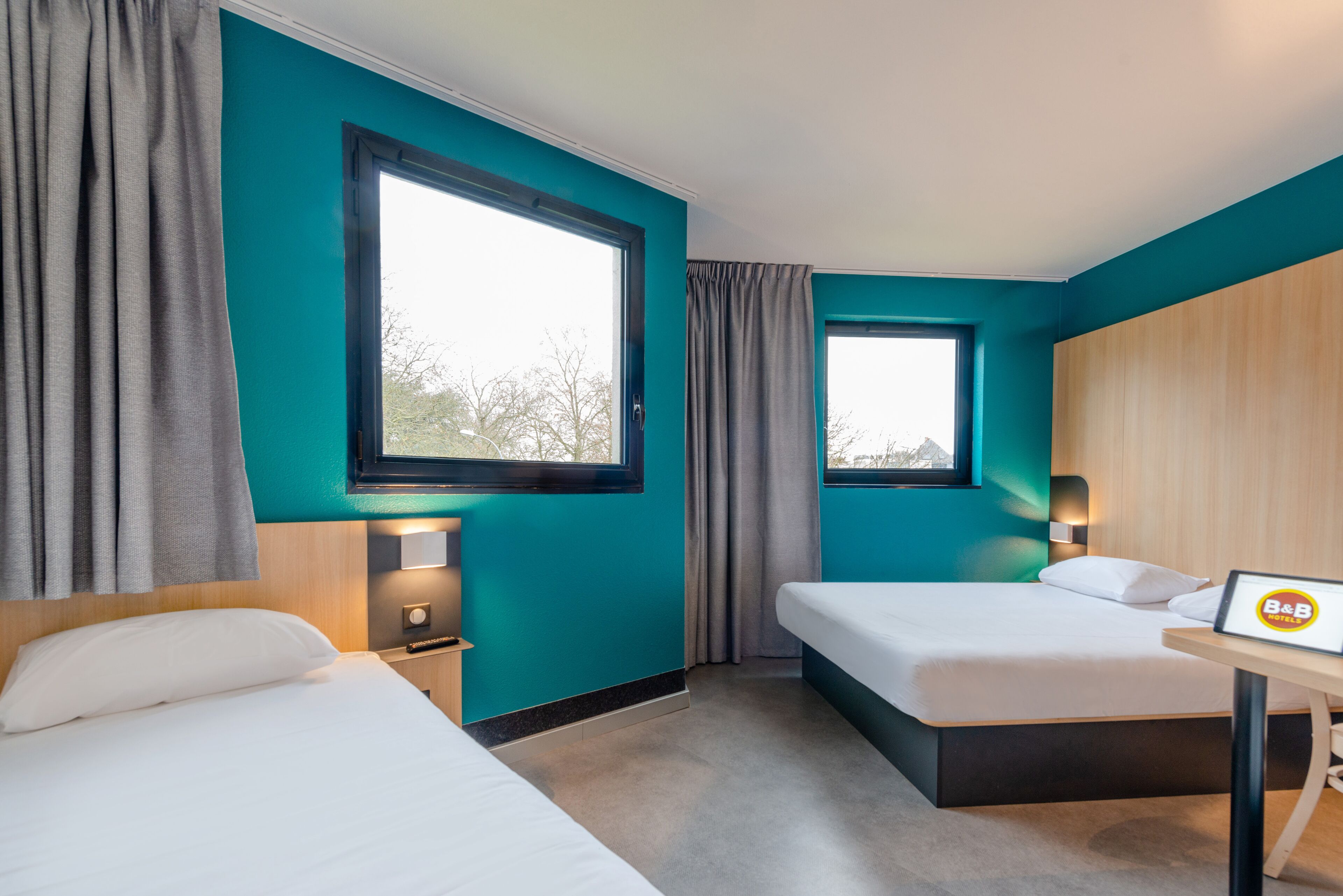 Egy szállodai szoba, melynek falai türkizkék színűek, két ágya van, egy íróasztal, rajta egy „B&B” felirattal, és egy ablak, melyen függöny van.