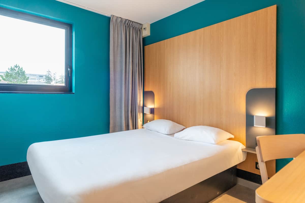 A hotel room with a bed, a window, a curtain, and a wall-mounted light fixture.
