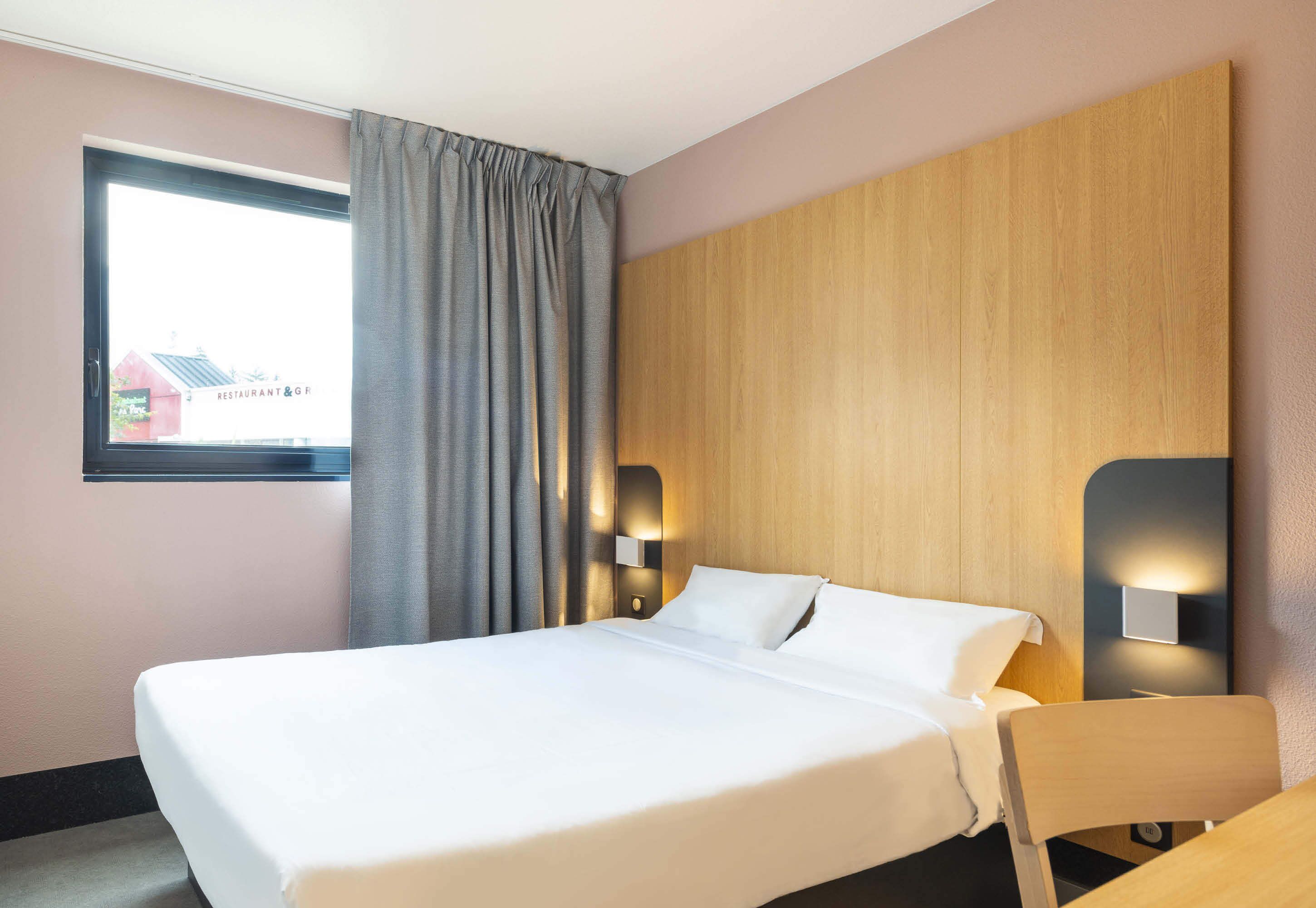 A hotel room with a large bed, a wooden headboard, a window with curtains, and wall-mounted lights.