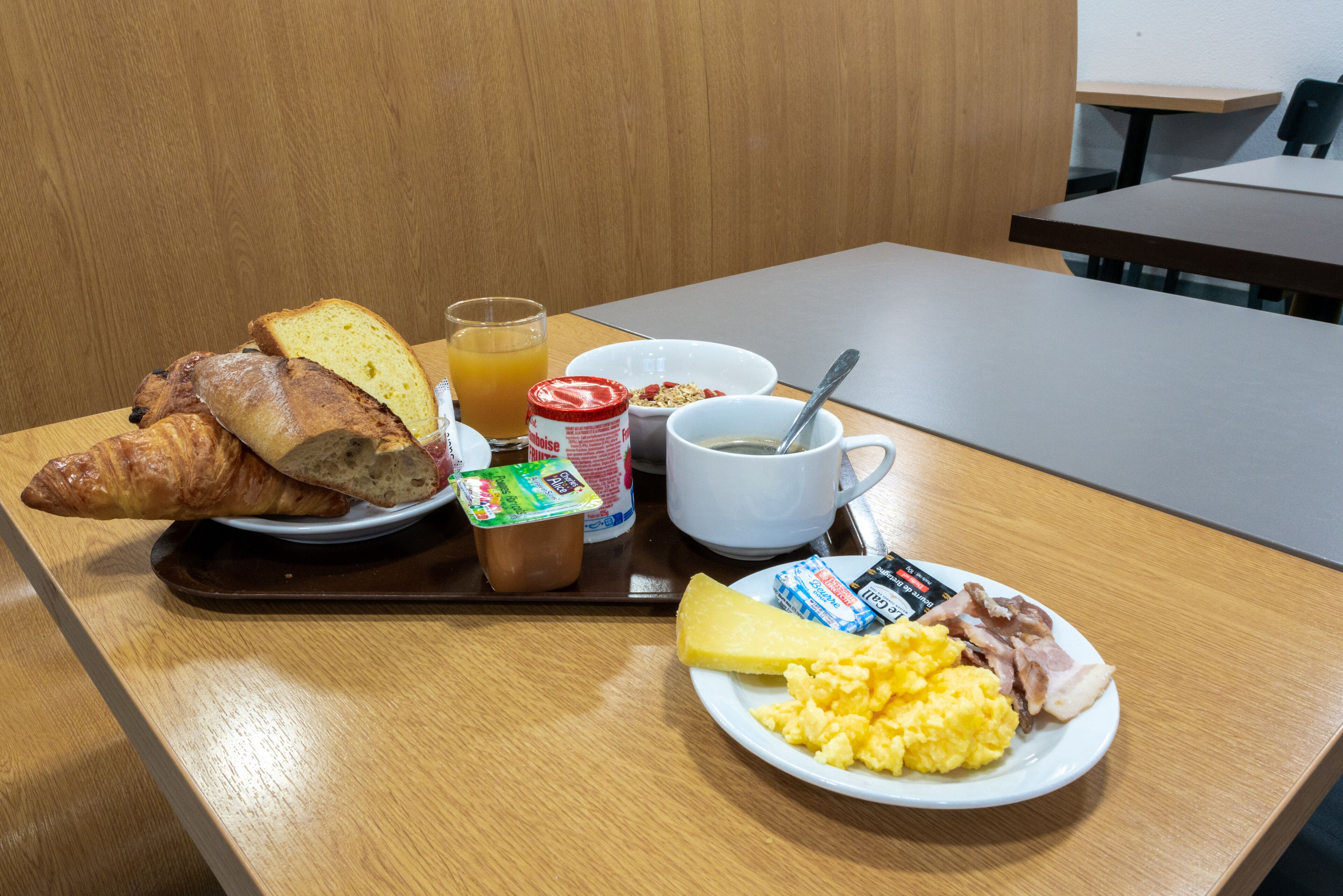 A breakfast tray with croissants, scrambled eggs, ham, cheese, a glass of orange juice, and a bowl of cereal.