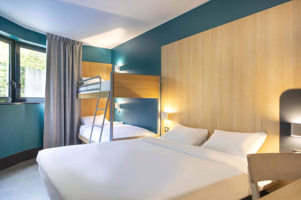 A hotel room with a bunk bed, a desk, a chair, a window with curtains, and a wall-mounted lamp.