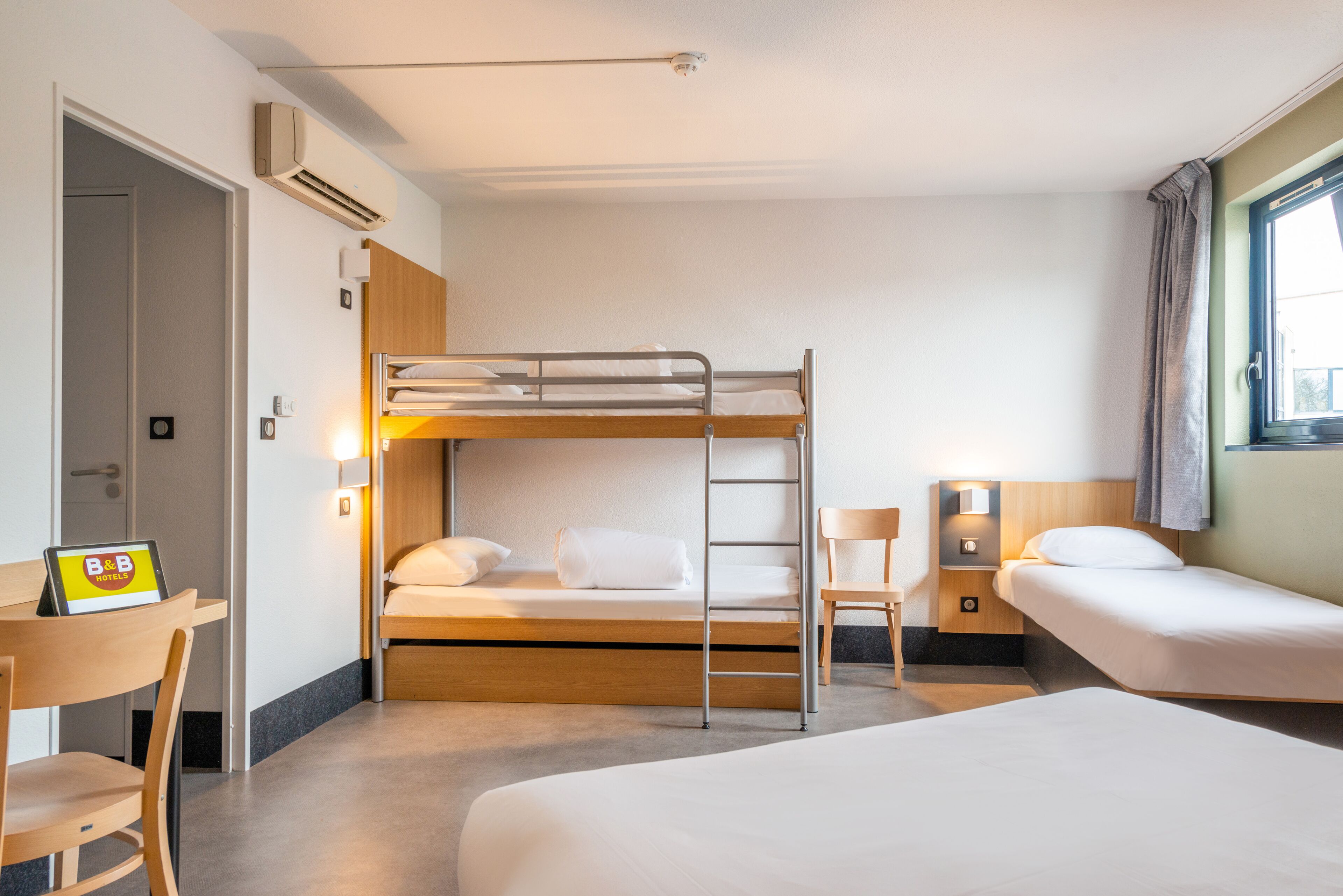 Une chambre d’hôtel avec un lit superposé, un bureau avec une chaise, une table de chevet et une fenêtre avec des rideaux.