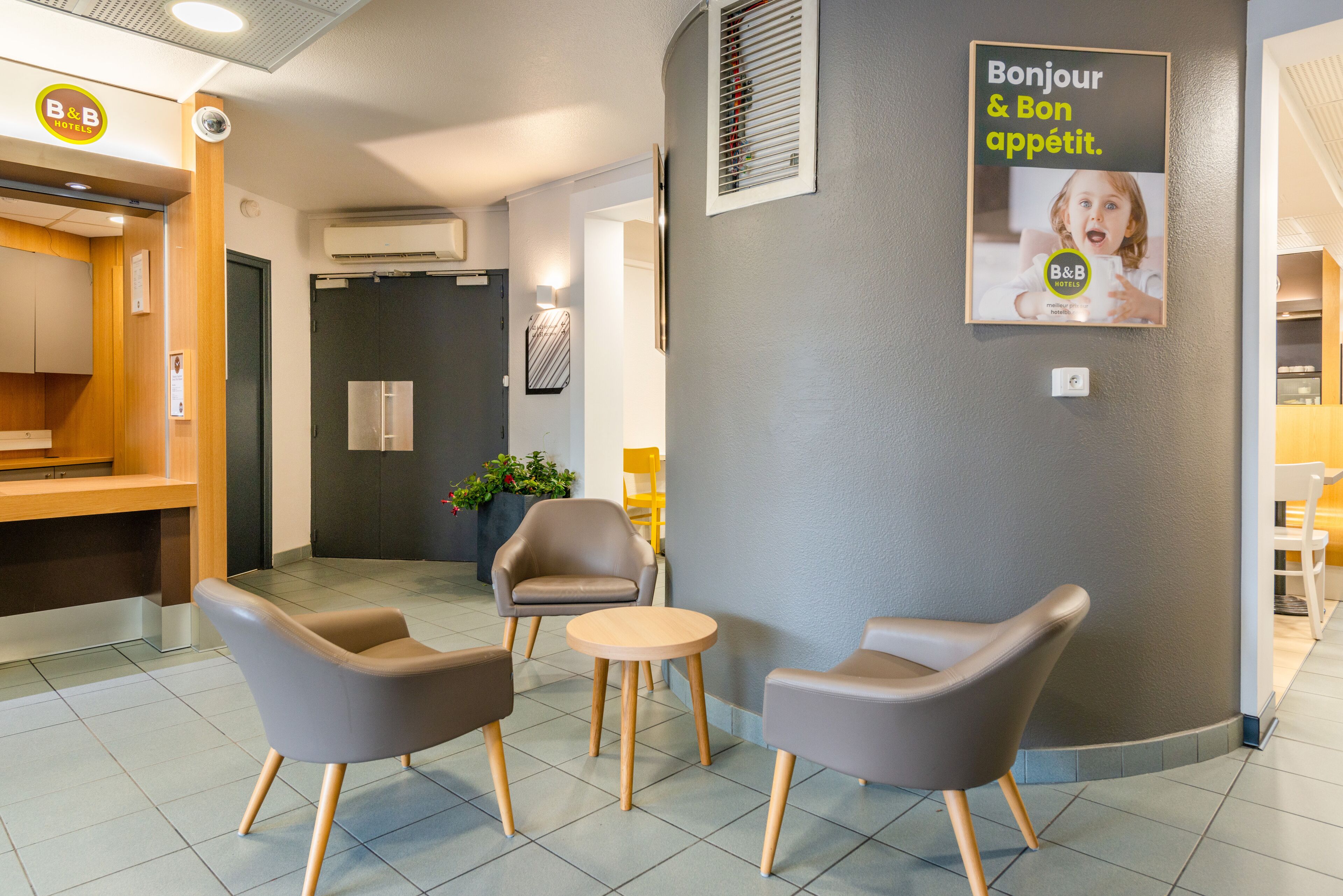 A hotel reception area with a modern design, featuring a reception desk, two armchairs, a small round table, and a wall-mounted sign with the hotel's logo.
