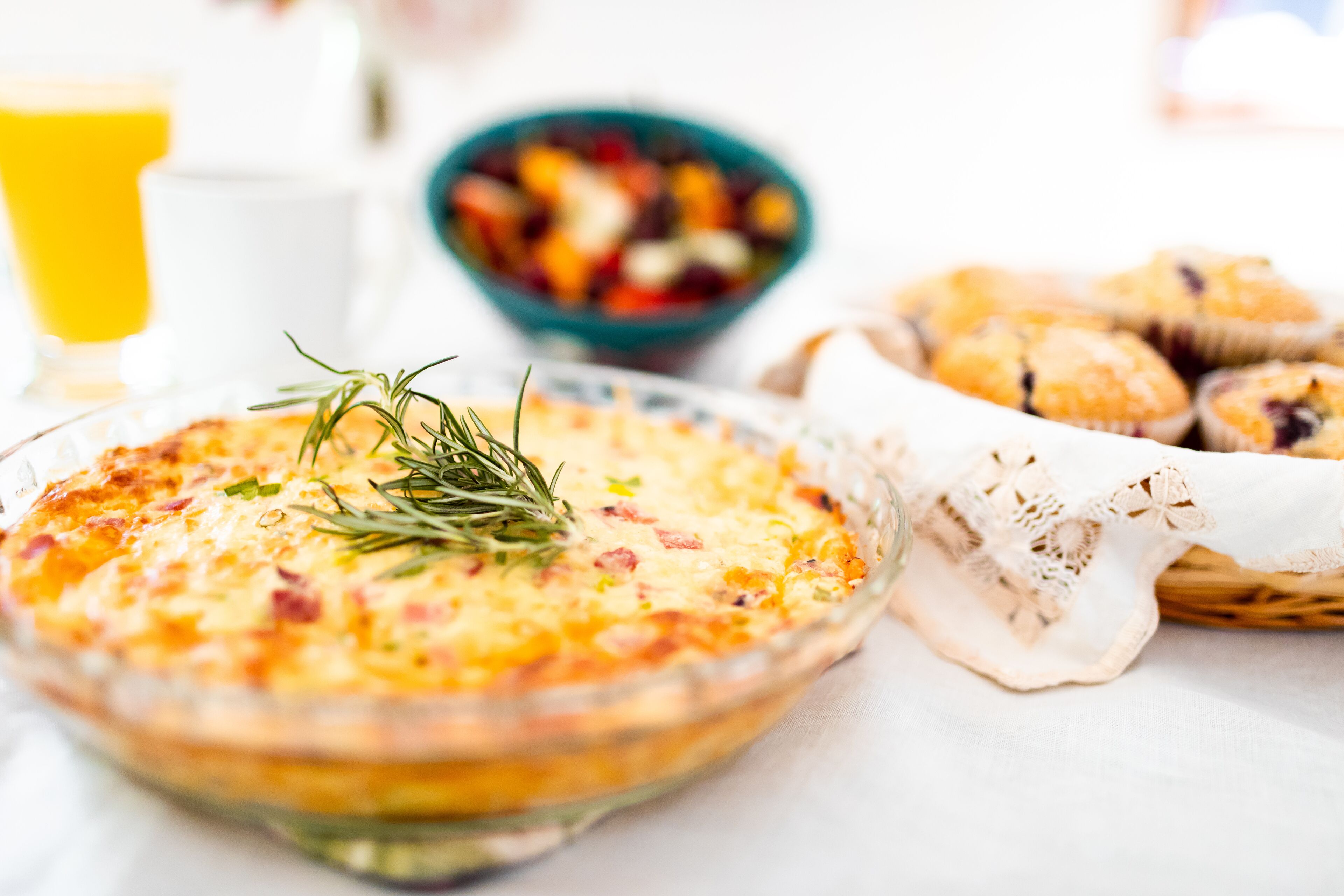A breakfast setting with a quiche, a bowl of fruit salad, and muffins on a table.