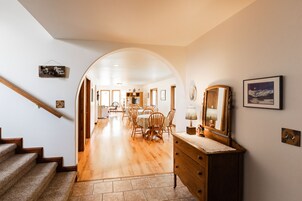 A spacious hallway with a wooden staircase, a dining area with wooden chairs and a table, and a wooden dresser with a mirror.