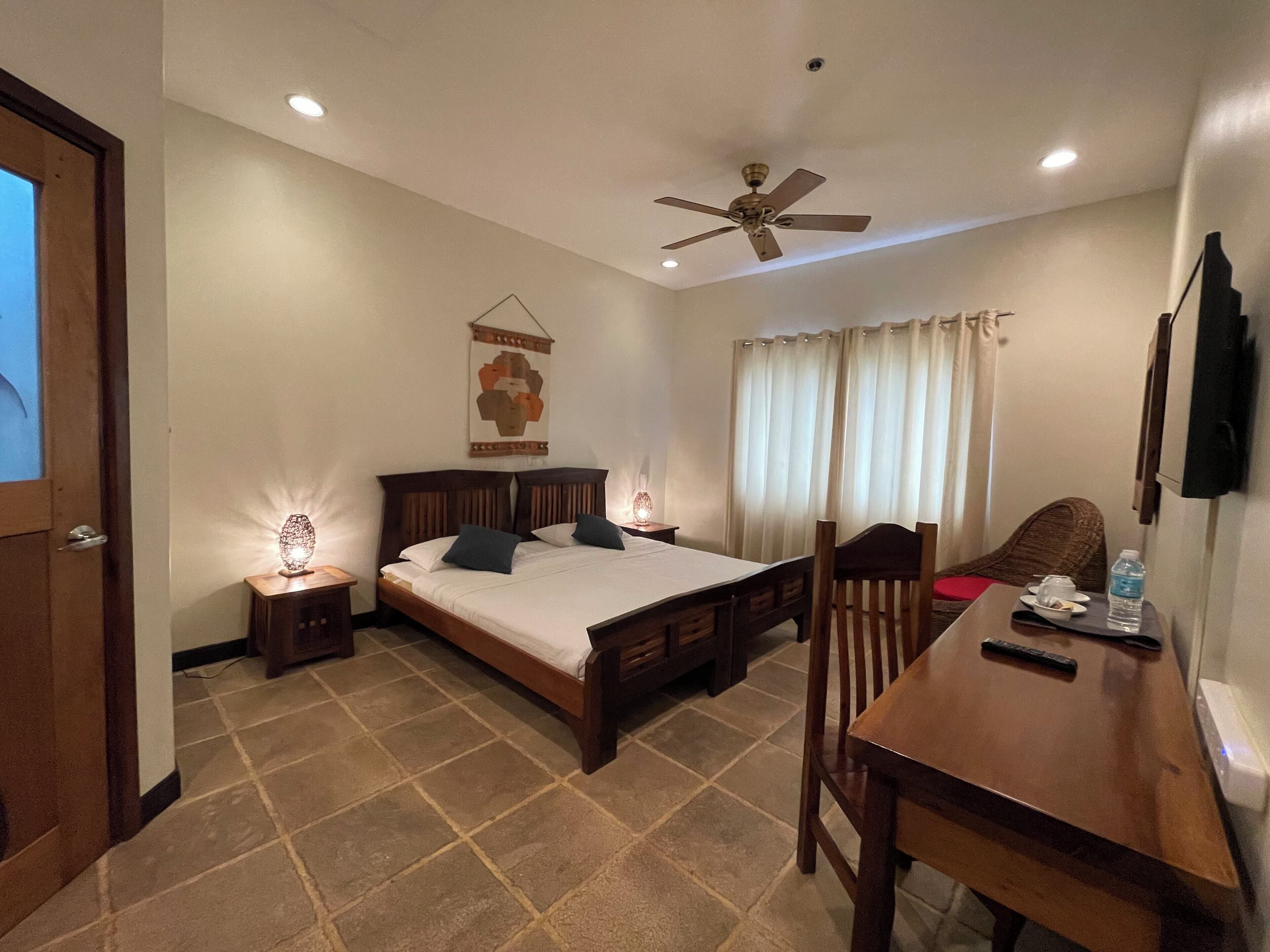 A hotel room with a bed, bedside tables, a desk, a chair, a ceiling fan, and a window with curtains.
