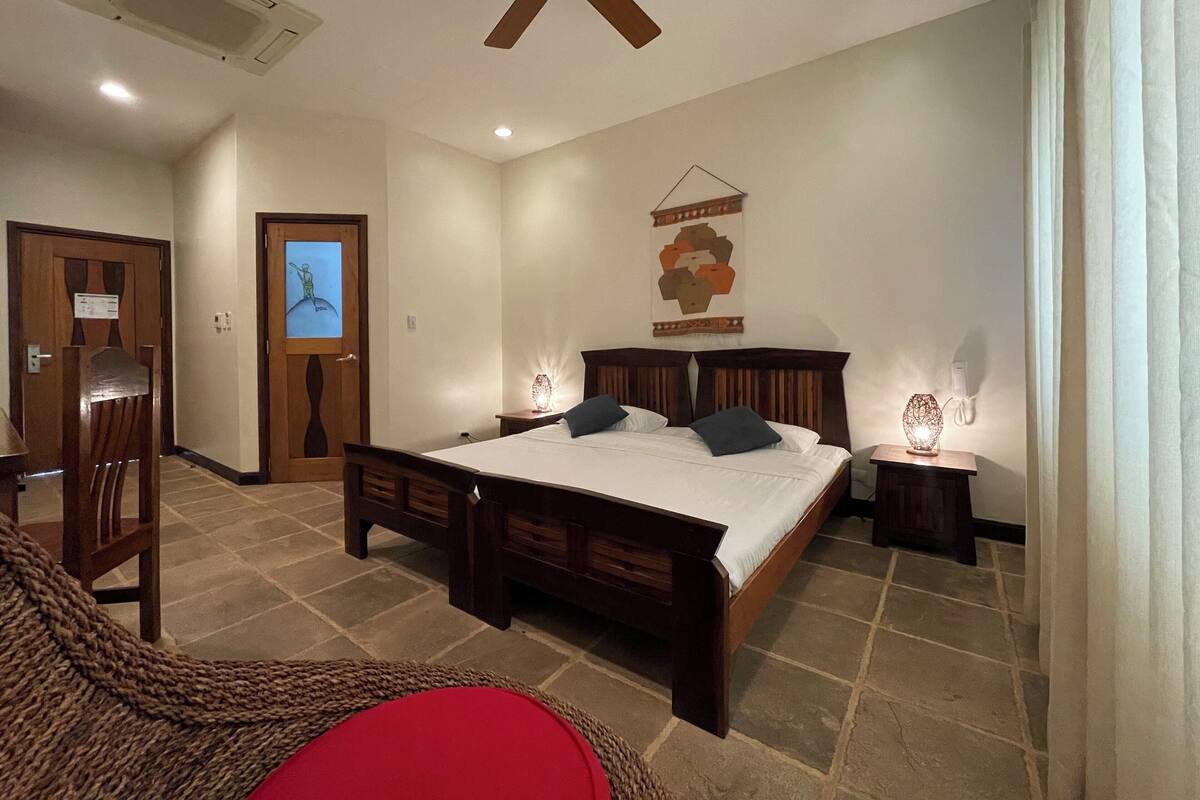 A bedroom with a large bed, a ceiling fan, a wall-mounted artwork, and a small table with a lamp.