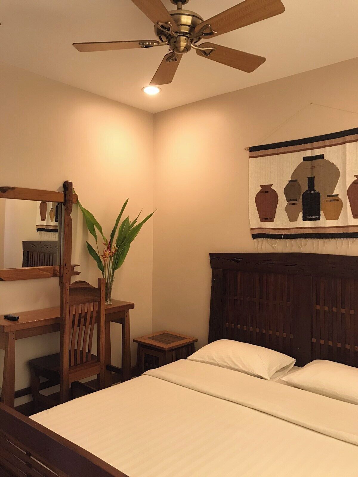A bedroom with a bed, wooden furniture, a ceiling fan, a small table, a chair, and a wall-mounted tapestry.
