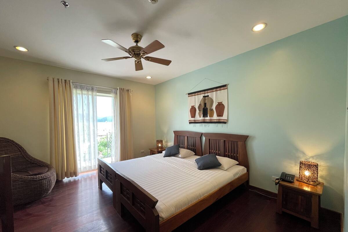 A bedroom with a large bed, a ceiling fan, a nightstand with a lamp, and a view of the outdoors through a window.