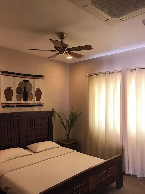 Una habitación de hotel con cama, ventilador de techo, planta en maceta y ventana con cortinas.