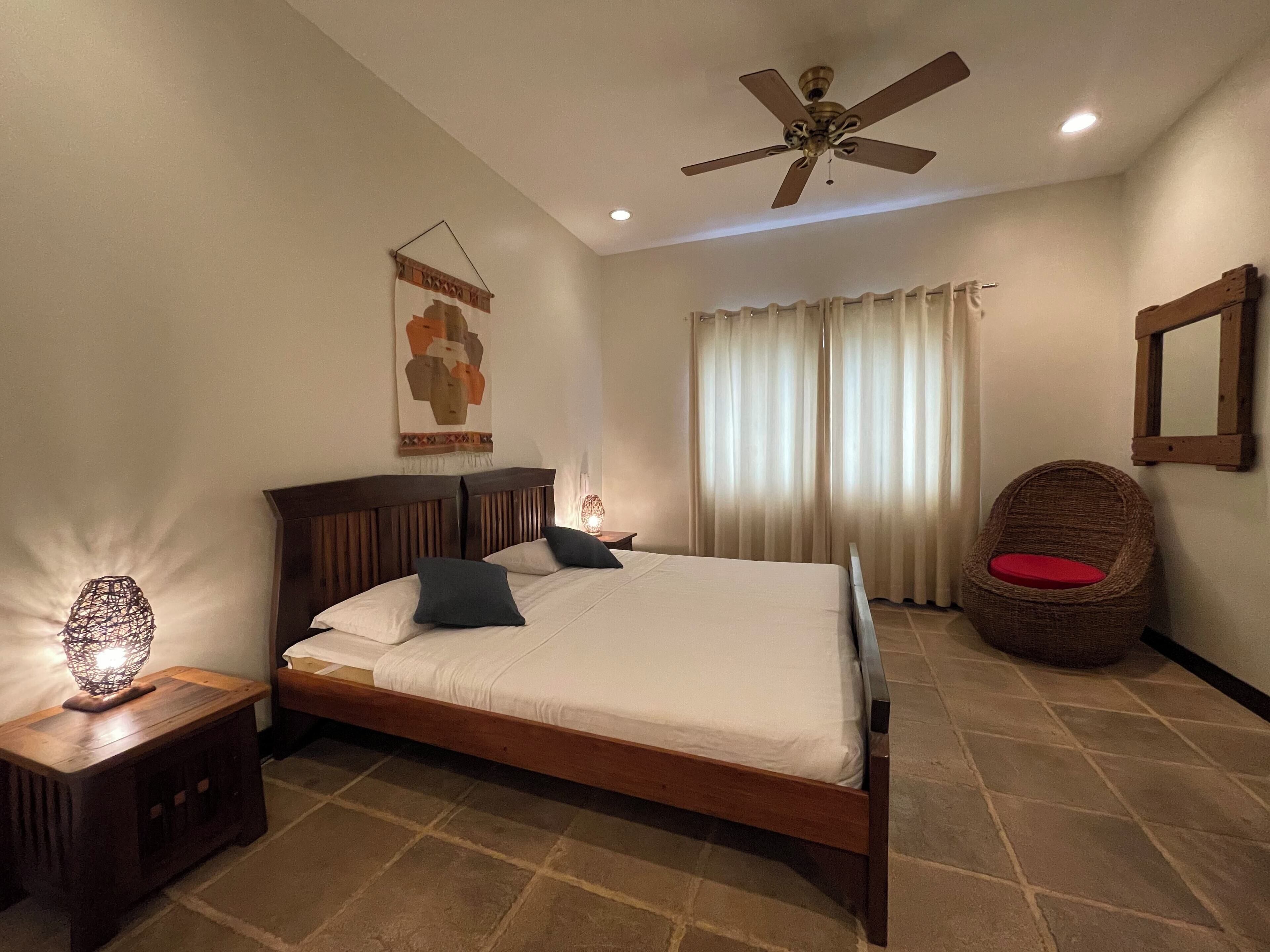 A bedroom with a wooden bed, a ceiling fan, a nightstand with a lamp, a wicker chair, and a window with curtains.