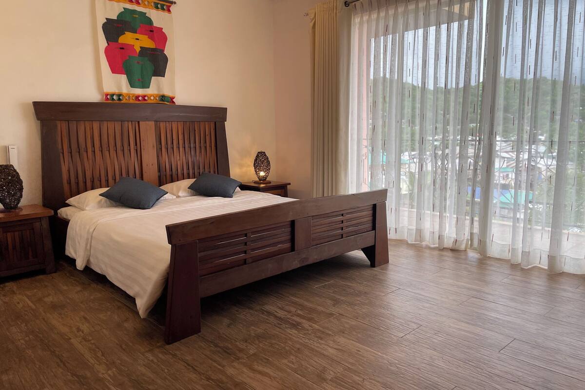 A bedroom with a wooden bed, two pillows, a nightstand with a lamp, and a large window with sheer curtains.