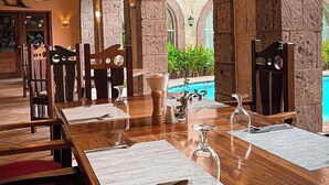 Un restaurante con una larga mesa de madera dispuesta para comer, arcos de piedra, luces colgantes y vistas a una piscina.