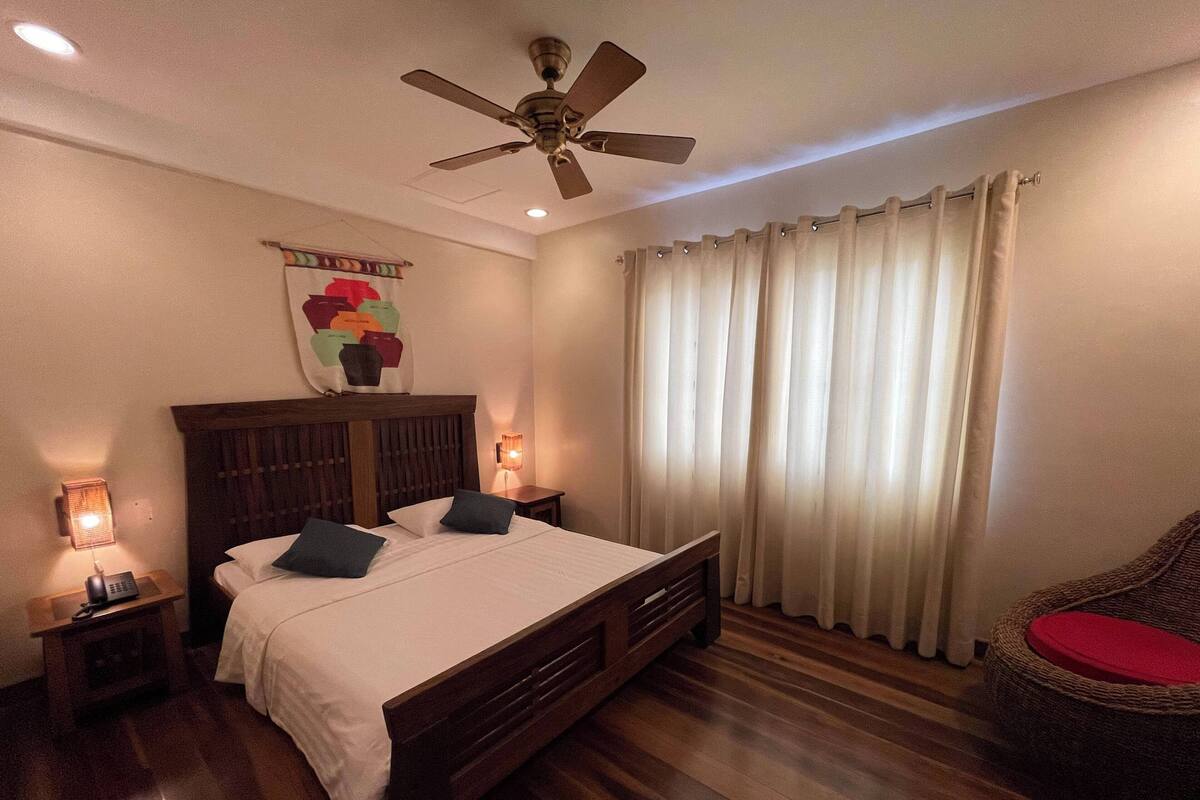 A bedroom with a wooden bed, a ceiling fan, a nightstand with a lamp, and a chair with a red cushion.
