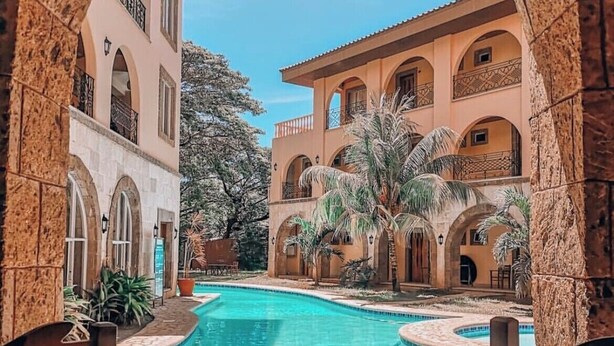 Zona de piscina con vista despejada a un edificio con múltiples balcones y arcos, rodeada de palmeras y un arco de piedra.