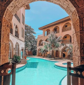 Zona de piscina con vista despejada a un edificio con múltiples balcones y arcos, rodeada de palmeras y un arco de piedra.