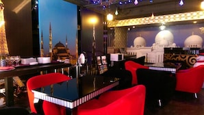 Un restaurante con sillas y mesas rojas, una pared decorativa con una mezquita y cúpula, y un fondo iluminado en azul.