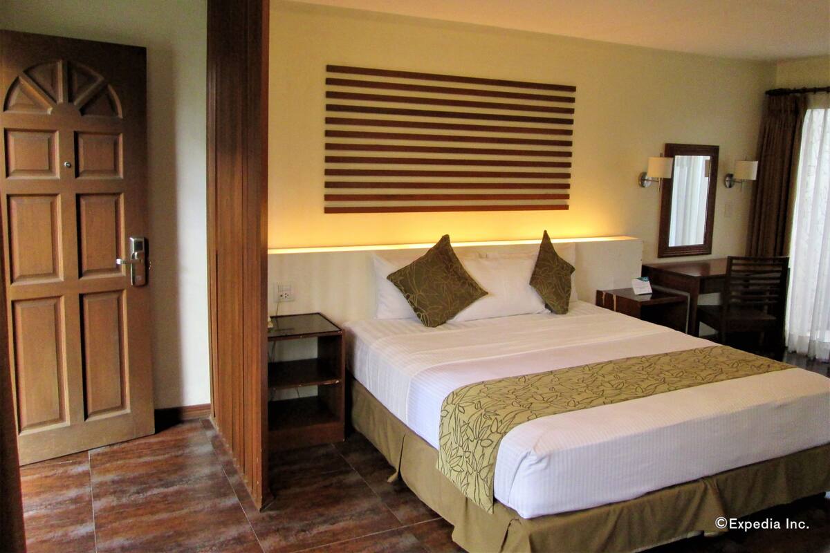 A hotel room with a large bed, a wooden door, a nightstand, a desk, and a mirror.