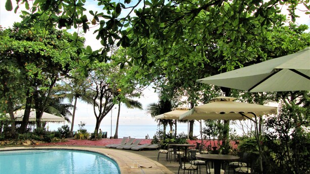 A poolside area with lounge chairs and umbrellas, surrounded by trees and overlooking a body of water. - Chali Beach Resort and Conference Center (Cagayan de Oro)