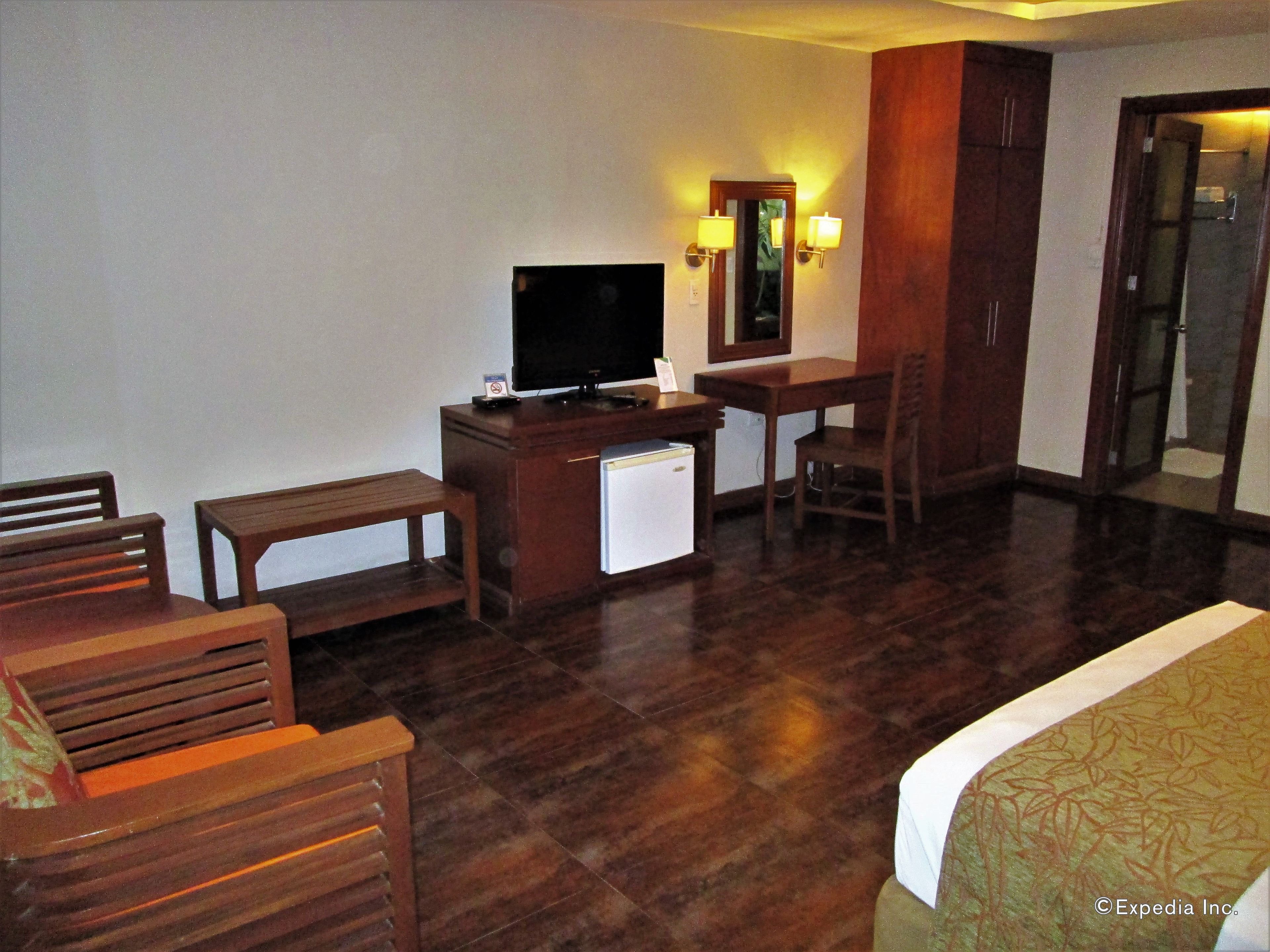 Habitación de hotel con un escritorio de madera, televisión, refrigerador pequeño, cama y espejo con apliques de pared.