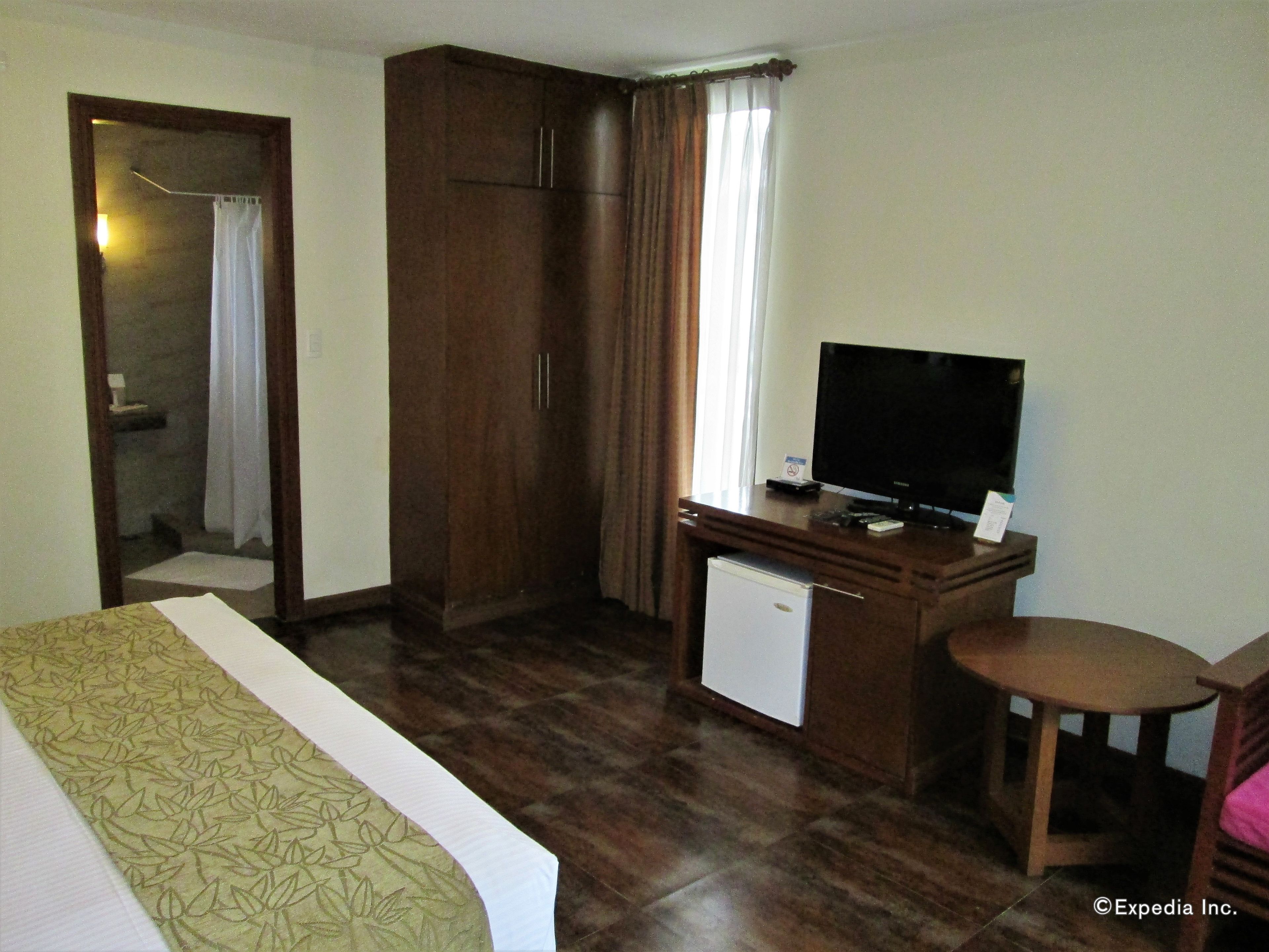 A hotel room with a bed, a desk with a television, a small refrigerator, and a wardrobe.