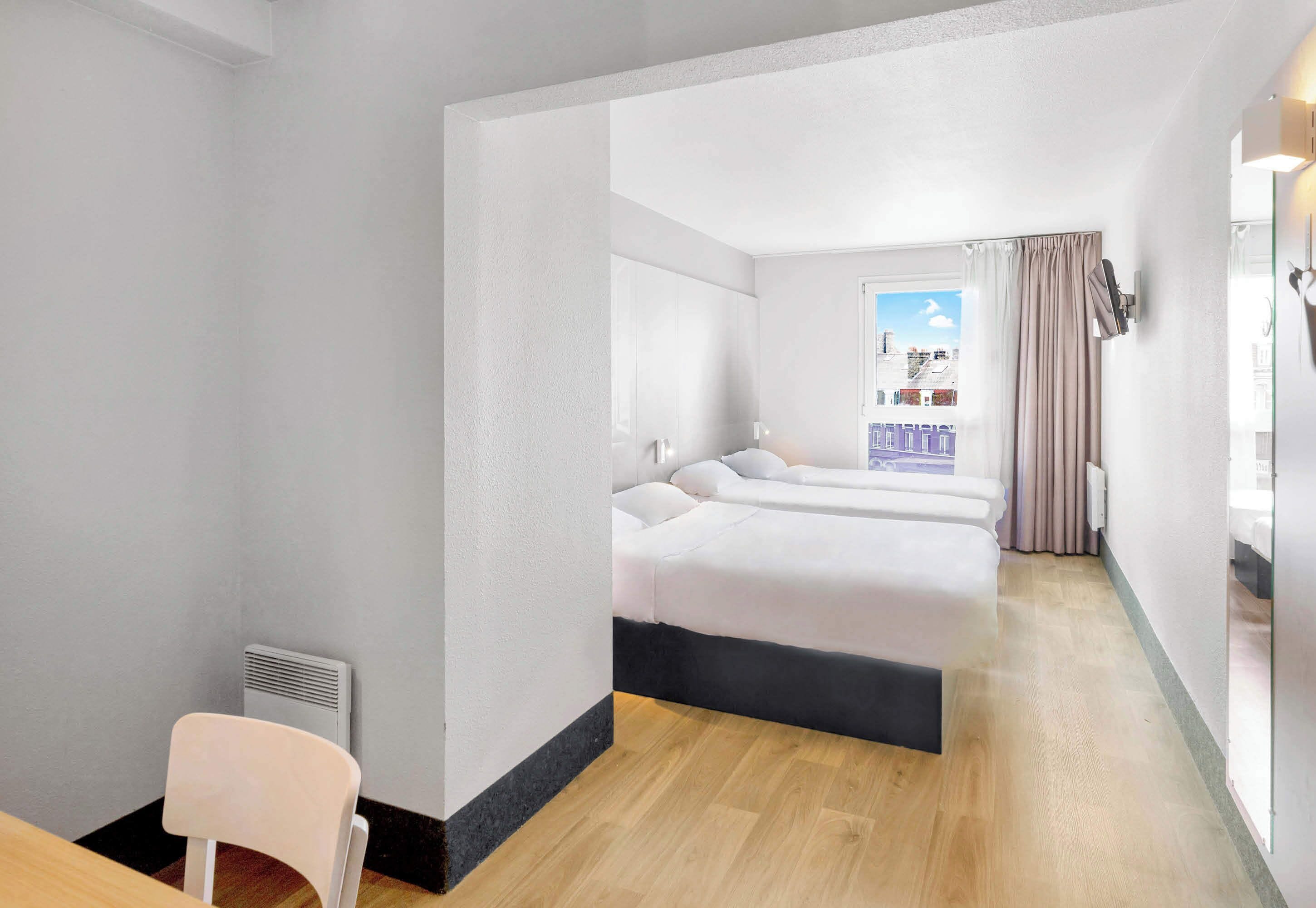 A hotel room with a bed, a chair, a window with curtains, and a view of buildings outside.