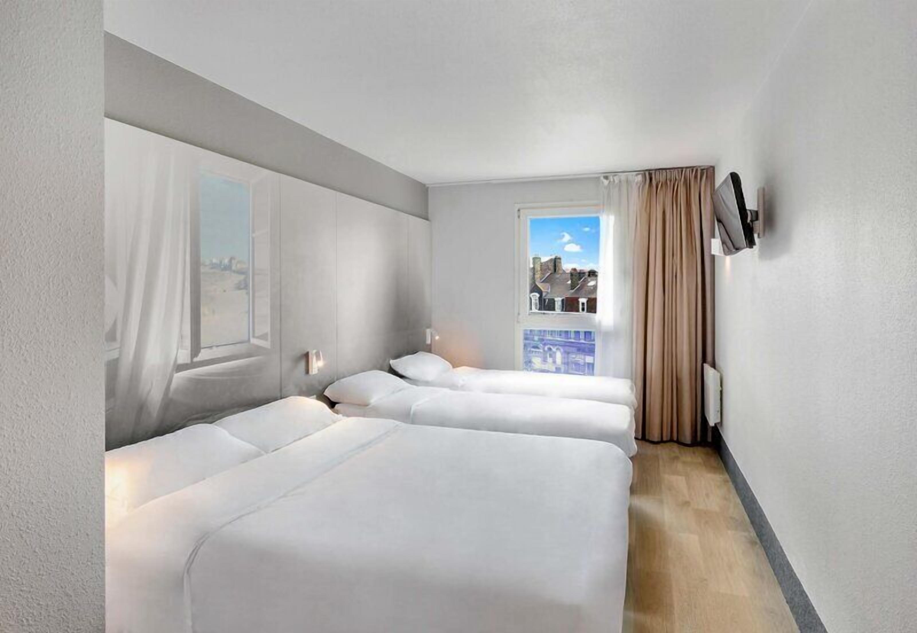 Habitación de hotel con dos camas, televisión, ventana con cortinas y vista a edificios.