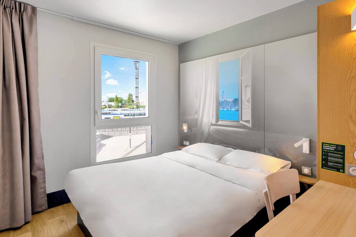 Hotellihuone, jossa on suuri sänky, puinen työpöytä, ikkuna, josta avautuu näkymä kaupunkiin, ja peili, joka heijastaa huonetta.