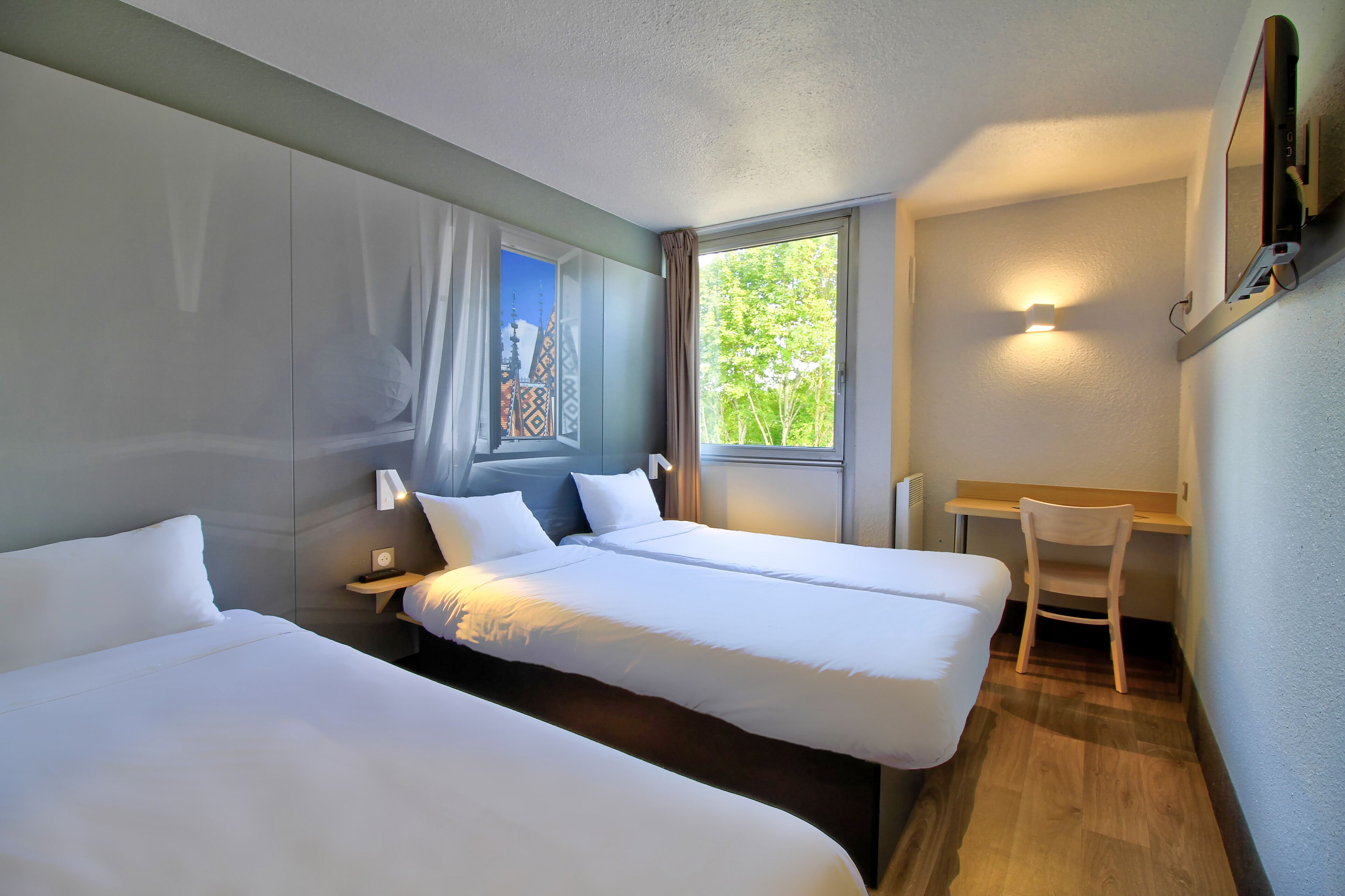 A hotel room with two single beds, a desk, a chair, and a window with a view of greenery.