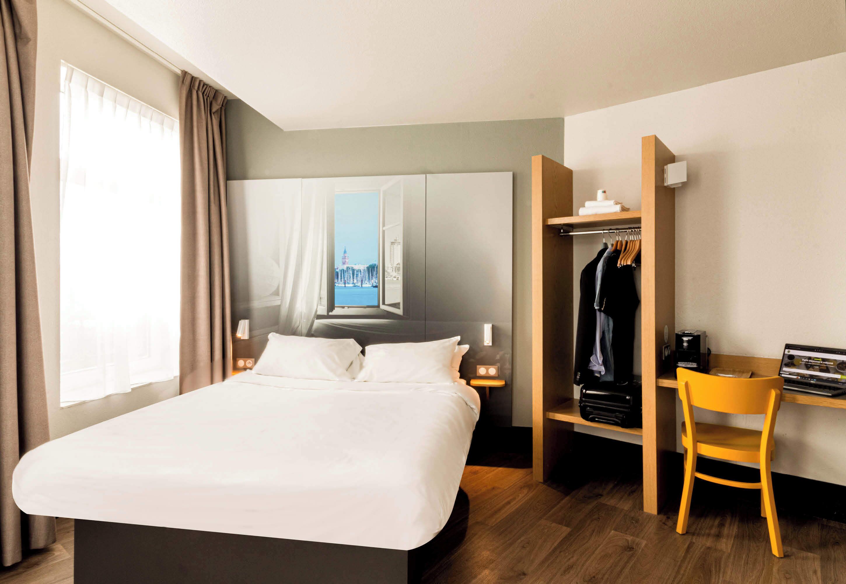 A hotel room with a bed, a desk with a chair, a wardrobe, and a window with curtains.