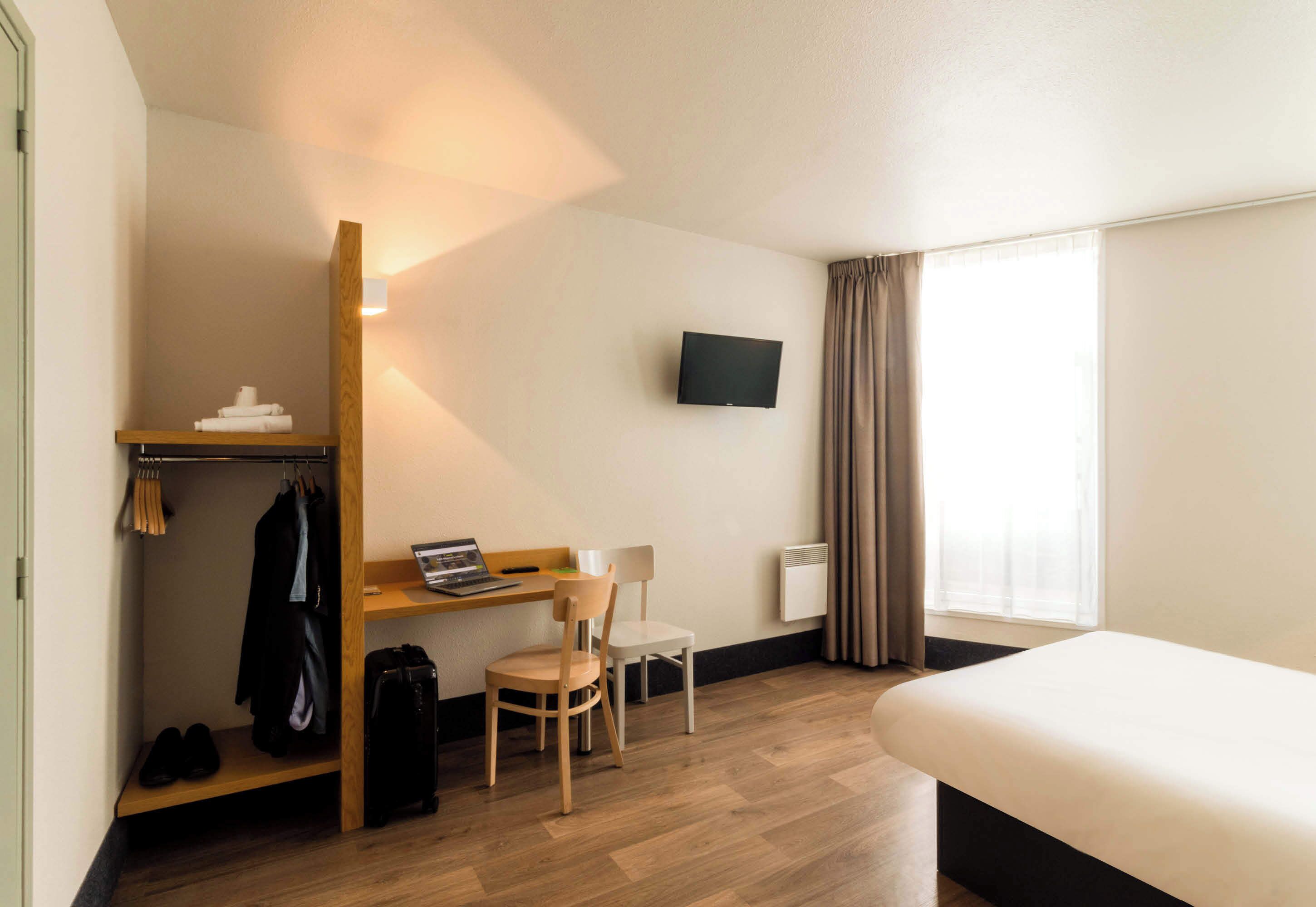 A hotel room with a bed, a desk with a laptop, a chair, a TV mounted on the wall, a coat rack, and a window with curtains.
