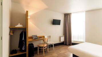 A hotel room with a bed, a desk with a laptop, a chair, a TV mounted on the wall, a coat rack, and a window with curtains.