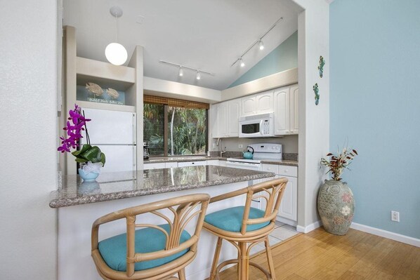 A modern kitchen with white cabinets, a granite countertop, and a central island with bar stools. - Wailea Ekahi Village, A Destination Residence (WAILEA)