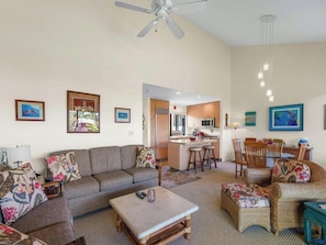 A living room with a sofa, ottoman, and chairs, a kitchen area with a dining table, and a ceiling fan.