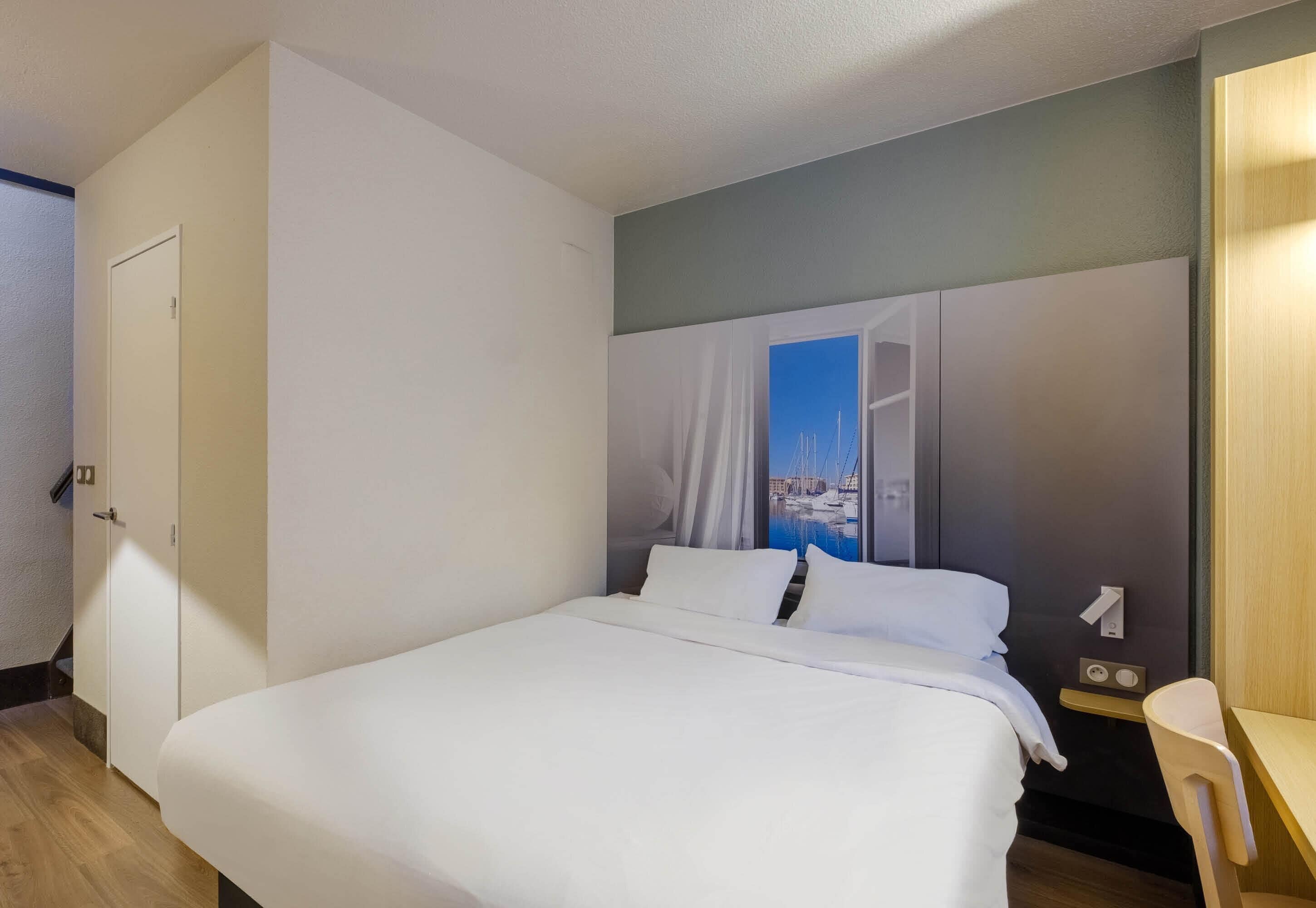 A hotel room with a bed, a desk, a chair, a window with a view of a marina, and a door.