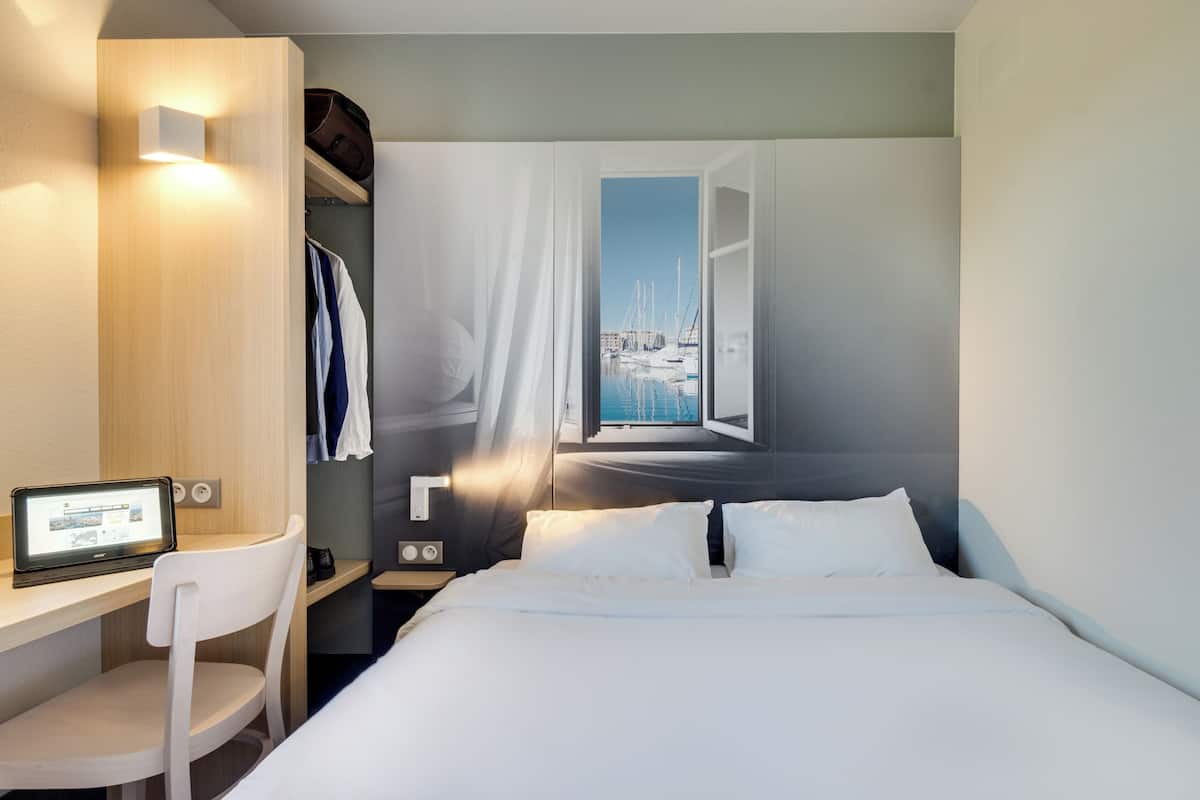 Une chambre d’hôtel moderne équipée d’un lit, d’un bureau avec un ordinateur portable, d’une chaise et offrant une vue sur une marina par une fenêtre ouverte.