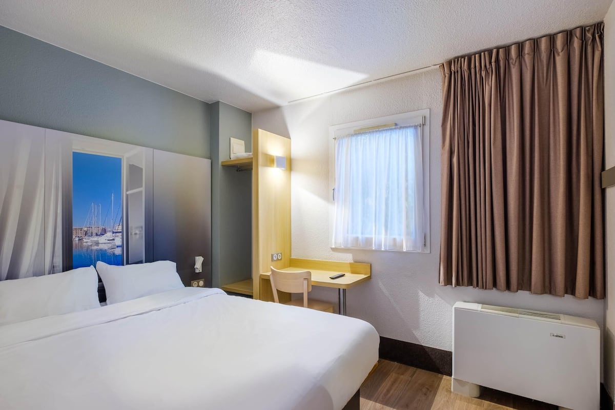 Une chambre d’hôtel avec un lit, un bureau, une chaise, une fenêtre avec des rideaux et un radiateur.